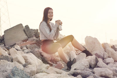 Young woman sitting on rock against sky