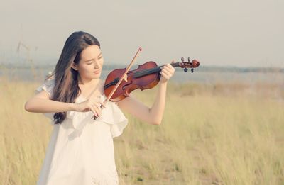 Young woman playing violin while standing on field against sky