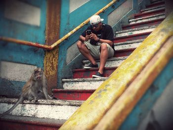 Young man photographing monkey on mobile phone while sitting on steps