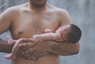 Midsection of shirtless father holding newborn son against wall