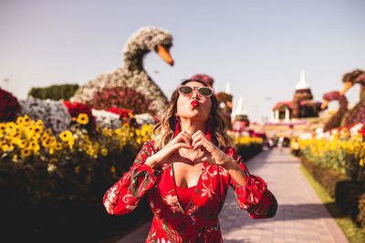 Woman wearing sunglasses making heart shape with hands while puckering lips outdoors