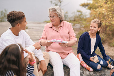 Happy grandmother giving gift to grandson while sitting with granddaughters in park during picnic