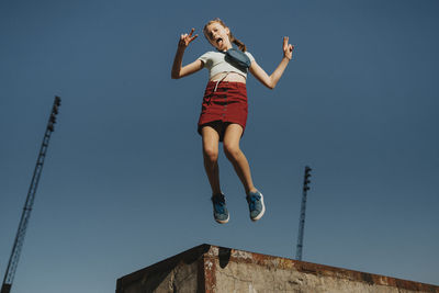 Carefree girl jumping in mid-air while doing peace sign against blue sky