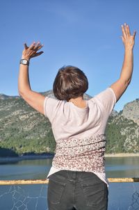 Rear view of woman with arms raised standing against lake and mountain