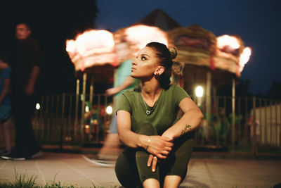 Thoughtful young woman looking away while sitting on footpath against sky at night