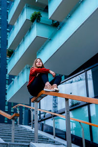 Low angle view of woman sitting on railing against building in city