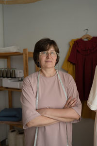 Self assured middle aged woman with tape measure crossing arms and looking at camera during work in atelier