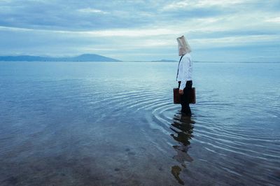 Man standing on lake with covered face against cloudy sky