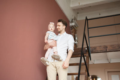 Male in white shirt holds baby girl standing on stair at their home.