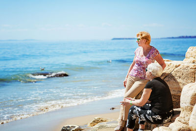 Two elderly women are happy to meet each other, sitting on rock on the seashore