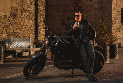 Man holding disposable cup while leaning on motorcycle parked against building