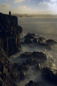 Long exposure image of man sitting on cliff against sky at sunset