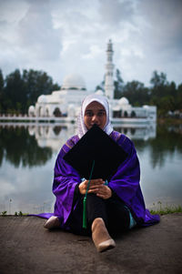 Woman holding umbrella while sitting by lake against sky