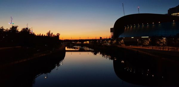 Silhouette buildings by river against sky during sunset