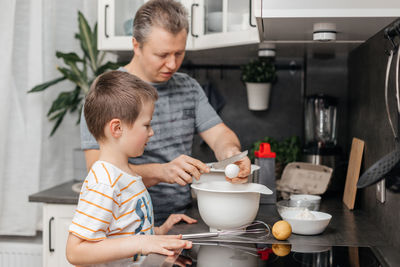 Dad and son are cooking in the kitchen. dad teaches to break the child's eggs and cook the dough.