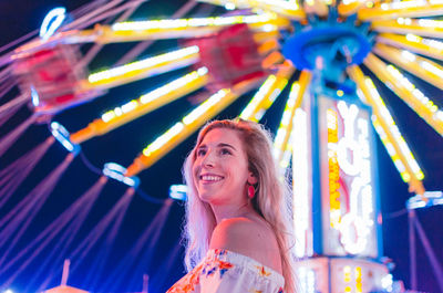 Low angle view of young woman standing against illuminated ferris wheel at amusement park