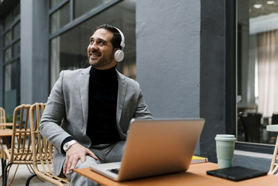 Smiling businessman with laptop and headphones looking away while sitting at cafe