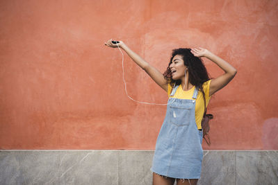 Cheerful woman listening to music on mobile phone while standing against wall