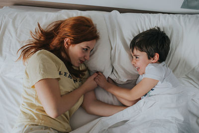 From above young woman and funny kid under duvet in pajamas looking at each other and lying on bed in bedroom