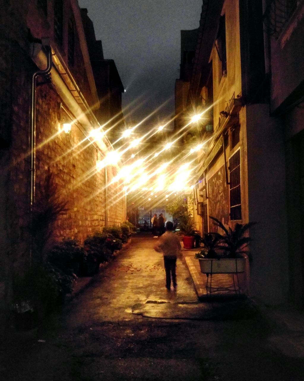 REAR VIEW OF MAN WALKING ON STREET AMIDST ILLUMINATED BUILDINGS