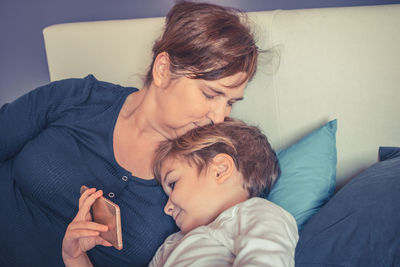 Grandmother kissing grandson holding mobile phone on bed