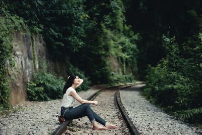 Side view of woman sitting on railroad track amidst trees in forest