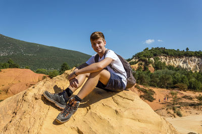 Portrait of smiling teenage boy sitting on cliff against blue sky