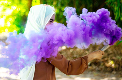 Side view of young woman holding purple distress flare while standing against trees