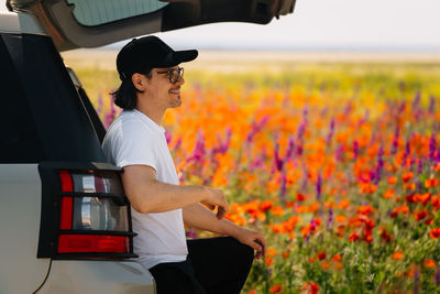 Side view of young man standing in suv car on field with poppy and wild flowers in summer