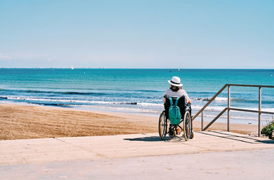 Back view of unrecognizable female traveler in wheelchair with backpack enjoying summer journey on beach near blue sea