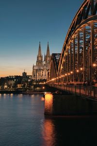 Illuminated bridge over river by buildings against sky at night