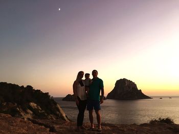 Family standing on cliff by sea against sky during sunset