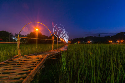 Boardwalk amidst rice field with light painting against sky at dusk