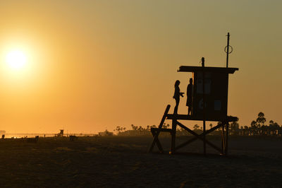 Silhouette lifeguards on hut at beach against orange sky