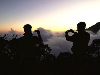 Rear view of silhouette friends photographing with smart phone while standing against sky during sunset