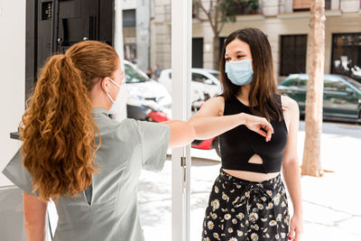 Professional staff in uniform and protective mask greeting female patient with gesture in entrance of modern spa salon during coronavirus pandemic