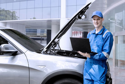 Portrait of smiling mechanic using laptop while standing against car