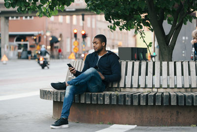 Young man using smart phone while sitting with legs crossed on bench in city