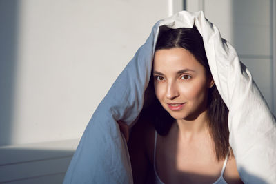 Woman in a white t-shirt is sit on the bed under the blanket in the room im morning