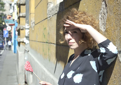 Young woman shielding eyes while holding lollipop by wall