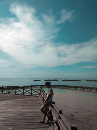 Man sitting on railing by sea against sky