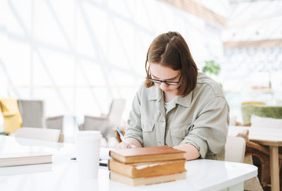 Young teenager girl college student in glasses doing homework with books at green library 