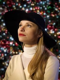 Close-up of thoughtful woman wearing hat against christmas lights at night