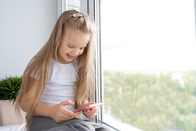 Smiling girl using mobile phone while sitting by window
