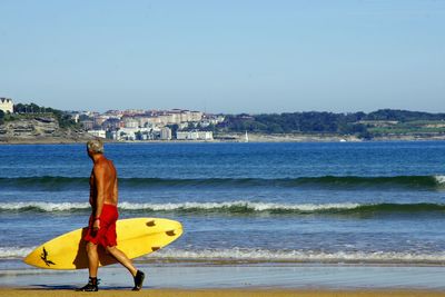 Shirtless man walking with surfboard at sea shore