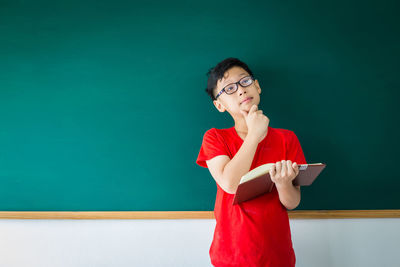 Thoughtful boy holding book while standing against blackboard