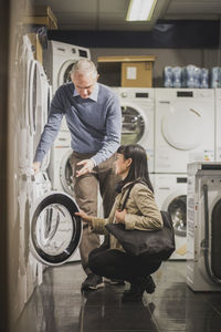 Mature owner explaining about washing machine to smiling female customer while standing in electronics store