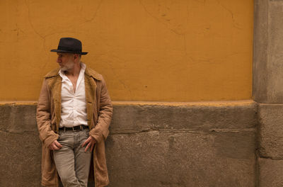 Portrait of adult man in hat and coat against yellow wall on street