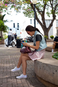 Side view of woman using phone while sitting on retaining wall