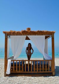 Woman wearing bikini standing at beach against clear blue sky